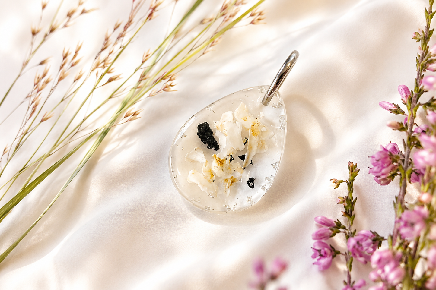 Minnessmycke med ingjuten aska i droppformat resin-hänge på ljus bakgrund med blommor.