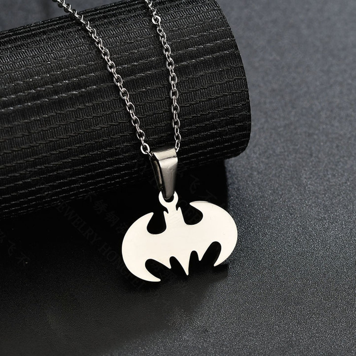 Ett Batman-inspirerat halsband i rostfritt stål med en guldfärgad och silverfärgad plätering, liggandes på en svart texturerad bakgrund.