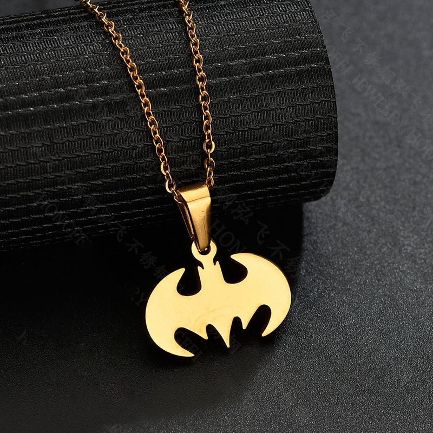 Ett Batman-inspirerat halsband i rostfritt stål med en guldfärgad och silverfärgad plätering, liggandes på en svart texturerad bakgrund.